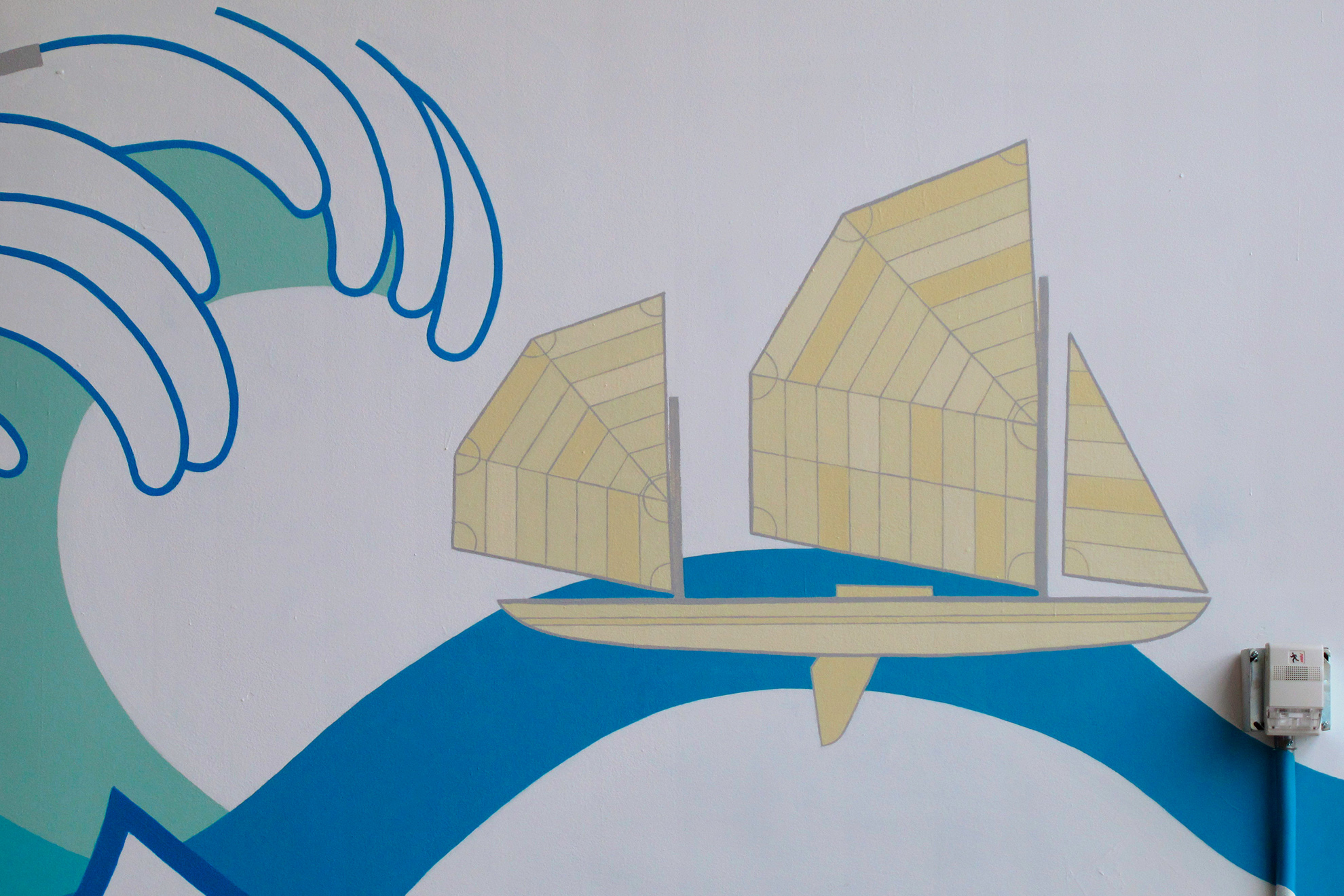 Zoom in Yakaboo sailing canoe in light mustardy yellows floating on a wave, 
                                                                    with the big frothy wave headed for it on its left.