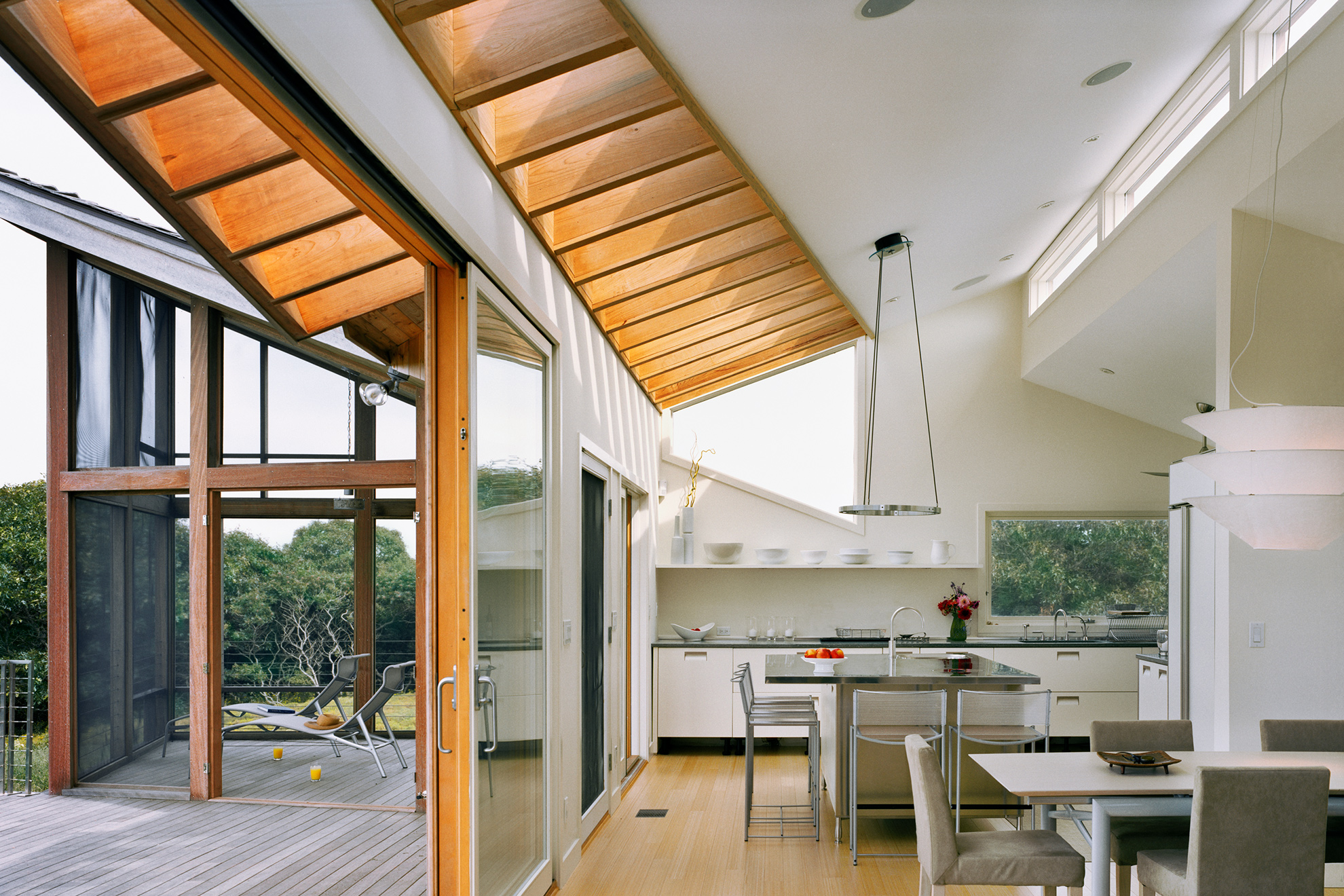 Wood exterior to white interior view along glass door line, with exposed wood rafters shown to continue from inside to outside, and many angled roof planes.
                                                                The enclosed wood screen porch is seen beyond on the left and the modern kitchen is seen beyond the living room table on the right.