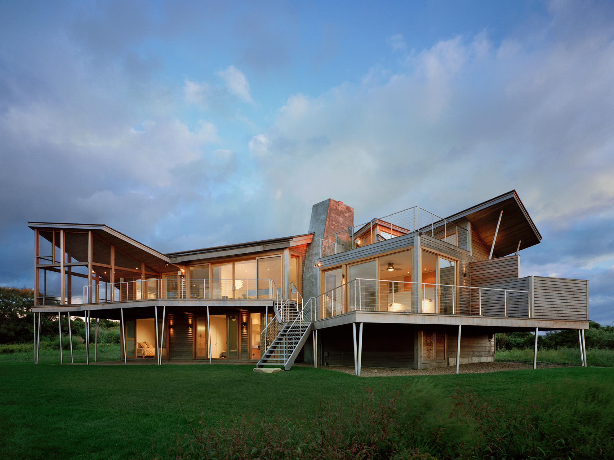 Explosive looking back side of vacation house, with roofs angled up in many directions, and clad in new horizontal cedar siding.
                                                                    A wood deck supported by very thin, round metal columns, and with a horizontal stainless steel cable railing and stairs coming down to the grass,
                                                                    floats above the lower floor the length of the house.