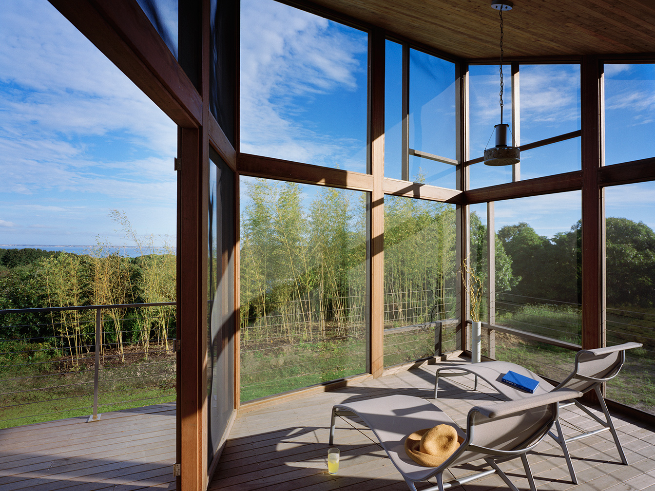 Tall, cedar wood framed outdoor screened porch looks out onto cedar deck with stainless steel cable railing and green vegetation in the foreground, with the ocean farther away in the background.