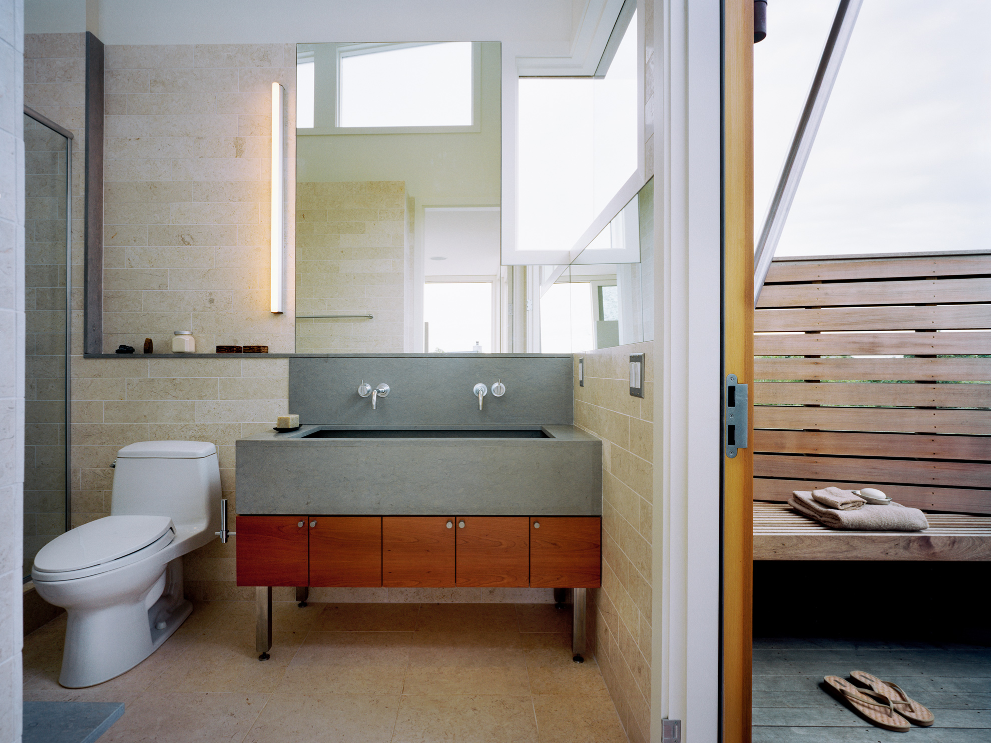 Interior to exterior view of a modern bathroom finished in an ivory-beige stone, and and outdoor shower area clad in new cedar siding.