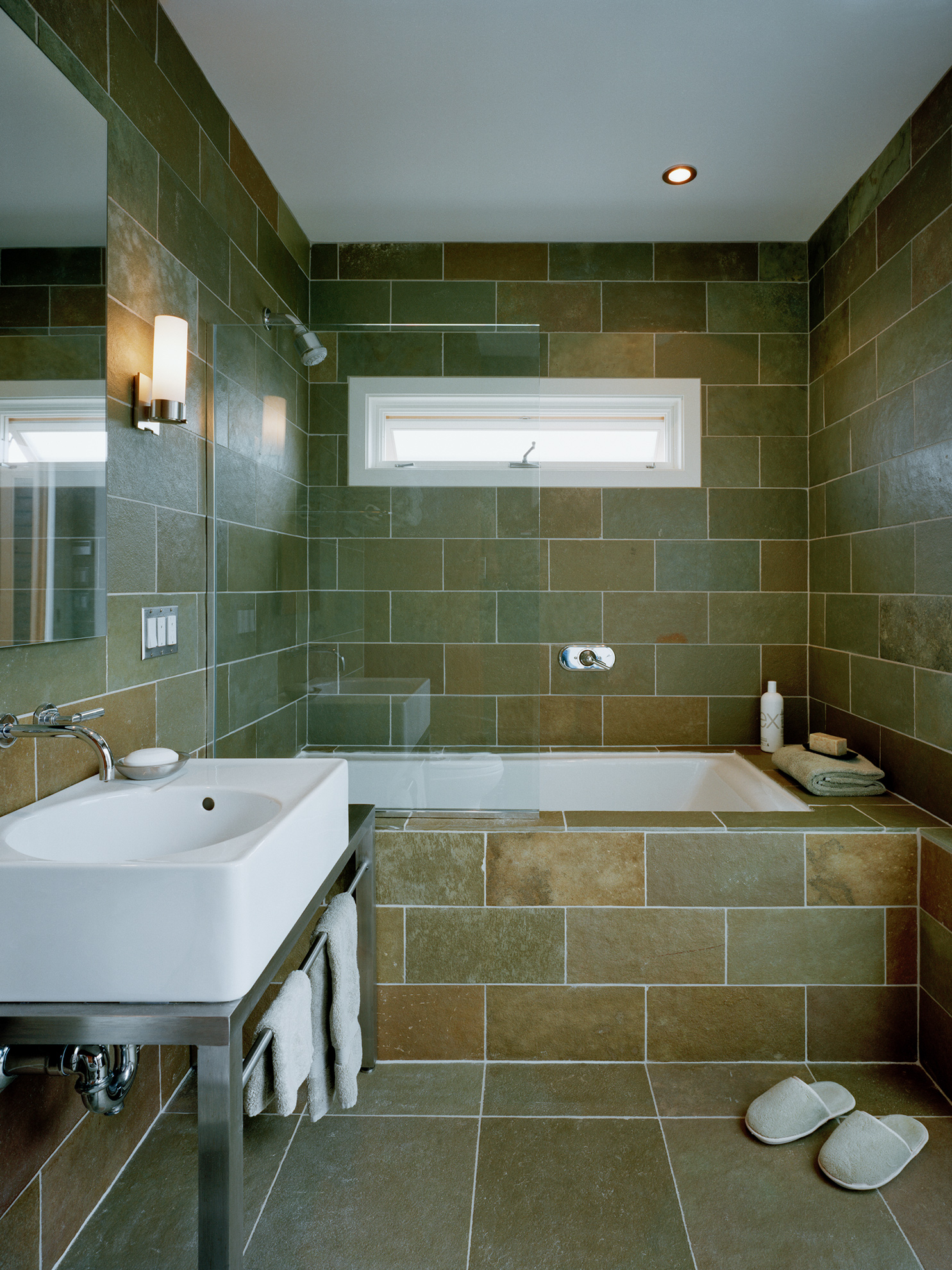 Modern bathroom interior with a greenish rusty stone tile around the walls and floor, and a long slim horizontal window above the shower/tub.