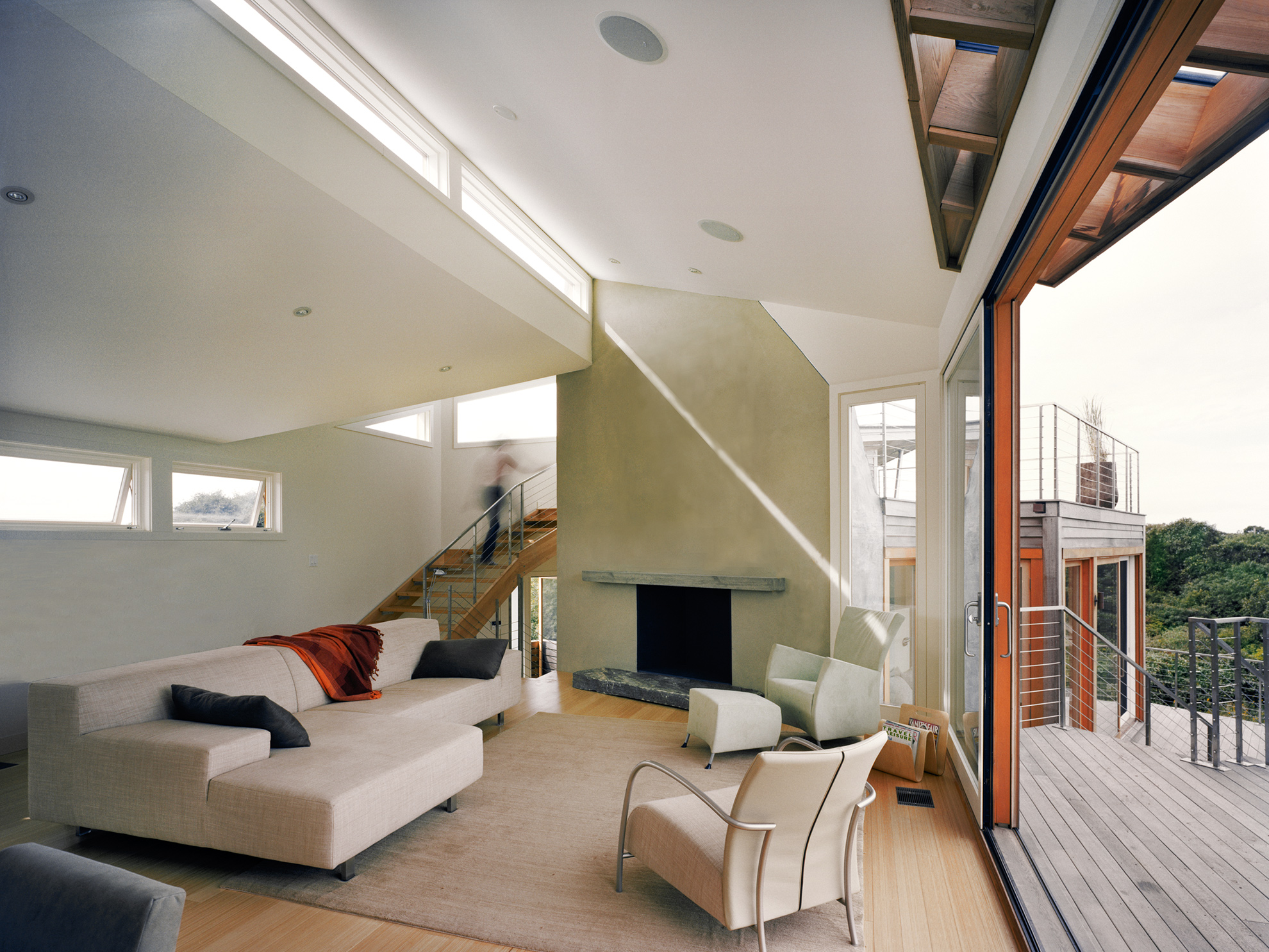 Living room interior with white furniture, a white concrete chimney, and exposed wood rafters in part of the angled ceiling/roof plane.
                                                                            Slivers of light are coming in through the long rows of clerestory windows, on two roof plane levels.