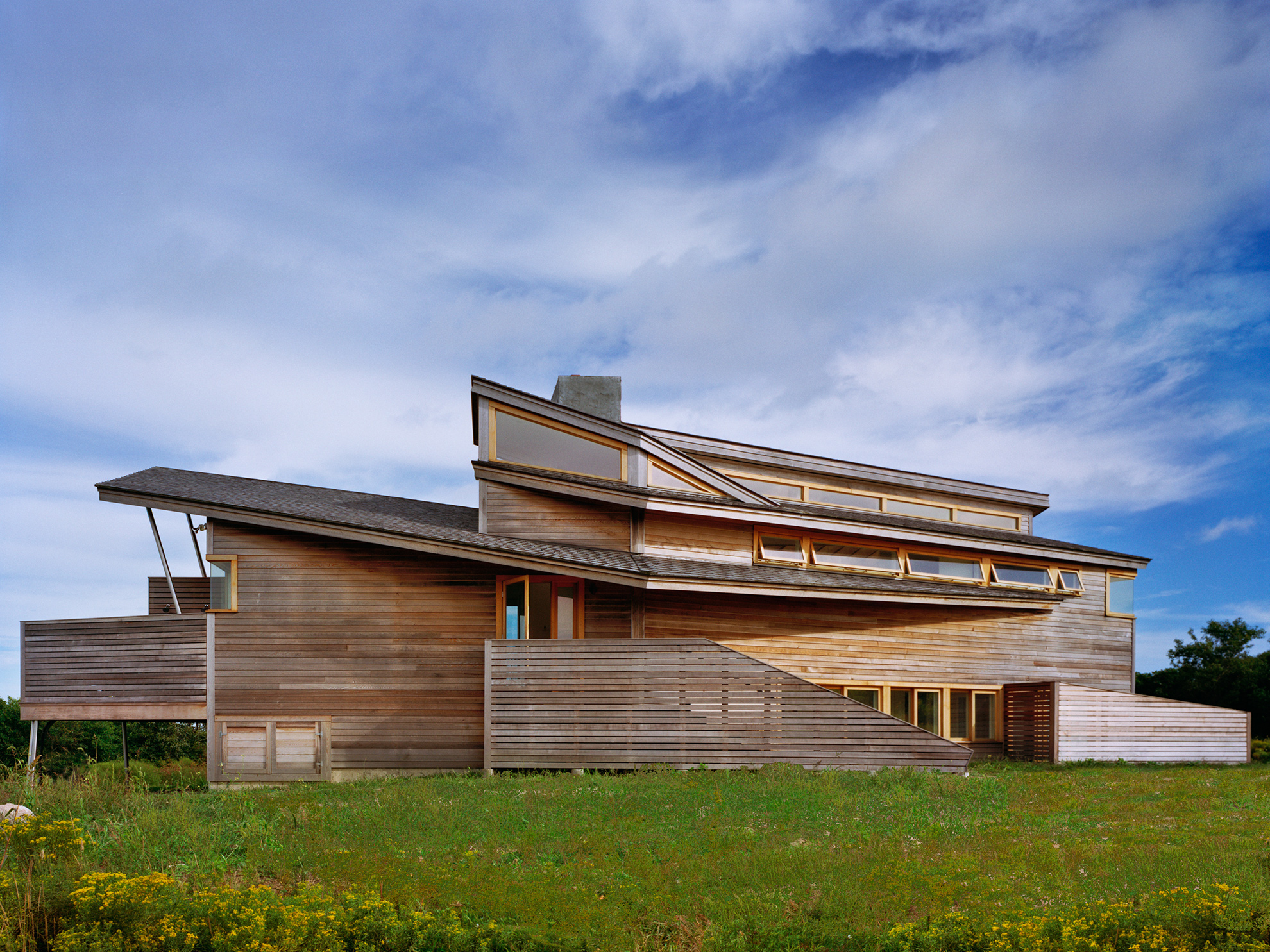A two story small longish house with layered angled walls and angled roofs, 
                                                                    clad in new horizontal cedar siding, sits in a grassy field against a big blue sky with clouds.
