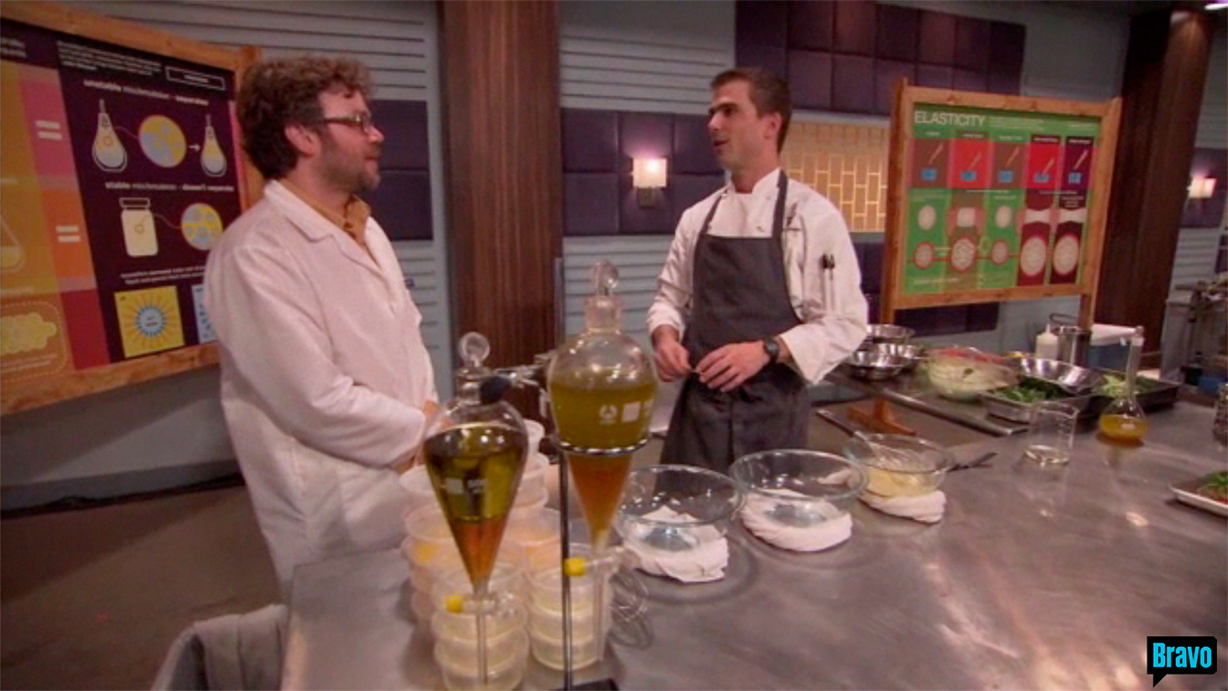 PhD student Augustine Urbas, dressed in a white lab coat, and Chef Hugh Acheson dressed in a white chef coat with a black apron on, 
                                                                talk in front of a stainless steel table with various beakers on it, on the set of Bravo TV&rsquo;s Top Chef Masters &ldquo;Blinded Me with Science&rdquo; episode. 
                                                                Behind them on the left is a large yellow and purple illustration on EMULSION, and on the right a large greenish illustration on ELASTICITY. 
