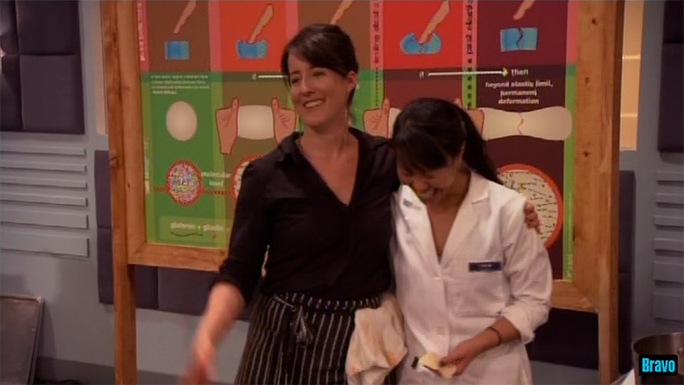Chef Naomi Pomeroy, dressed in a black button down shirt, hugs PhD student Carlin Hsueh, dressed in a white lab coat, while they both laugh in front of the greenish illustration on ELASTICITY,
                    on the set of Bravo TV&rsquo;s Top Chef Masters &ldquo;Blinded Me with Science&rdquo; episode.