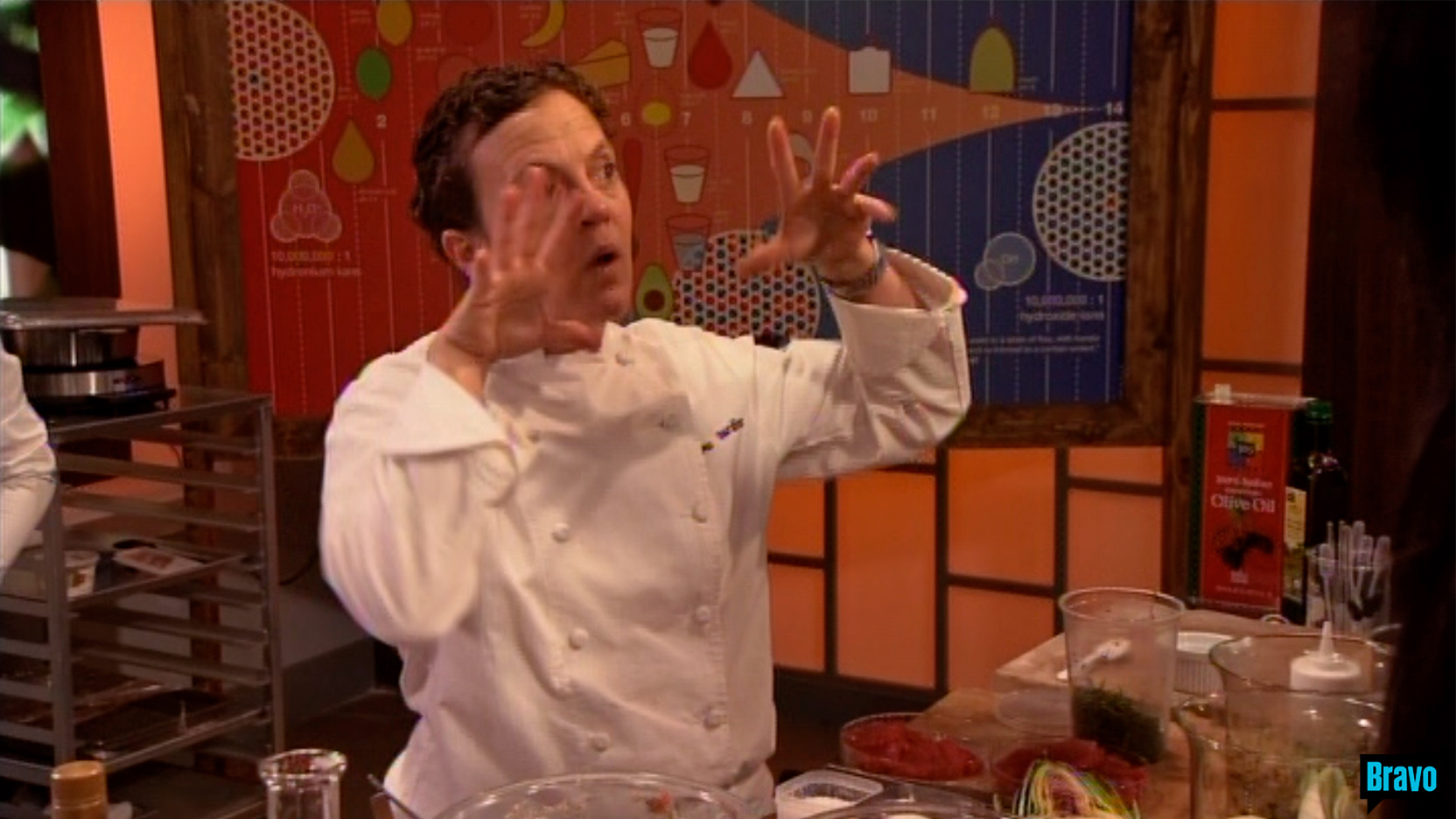 Chef Traci Des Jardins, dressed in a white chef outfit motions with hands up at face level,
                                                                             in front of a large ACIDITY illustration framed in stained pine is behind them, 
                                                                             on the set of Bravo TV&rsquo;s Top Chef Masters &ldquo;Blinded Me with Science&rdquo; episode.