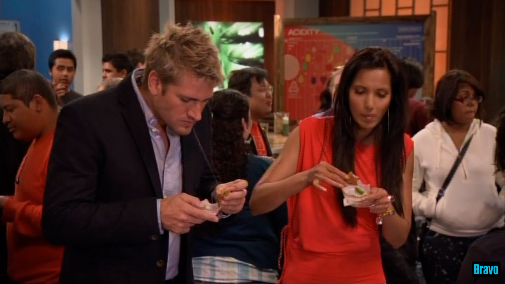 Curtis Stone in an open collar blue shirt with a navy jacket on and Padma Lakshmi in a bright orange dress taste some food.
                                                                                    There are a lot of people and high school students roaming behind them, and on the right can be seen the large red and blue ACIDITY poster,
                                                                                    on the set of Bravo TV&rsquo;s Top Chef Masters &ldquo;Blinded Me with Science&rdquo; episode.