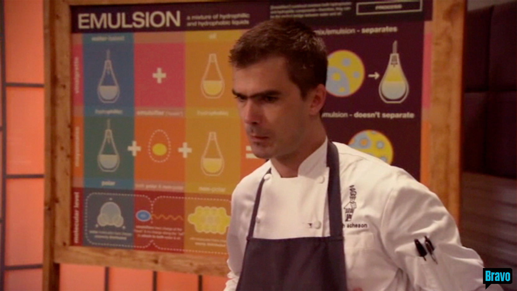 Zoom in on Chef Hugh Acheson&rsquo;s upper body, dressed in a white chef coat with a black apron and two pens in his left arm&rsquo;s pocket, 
                                                                                head looking down, concerned, standing in front of the large EMULSION illustration framed in stained pine, 
                                                                                on the set of Bravo TV&rsquo;s Top Chef Masters &ldquo;Blinded Me with Science&rdquo; episode.