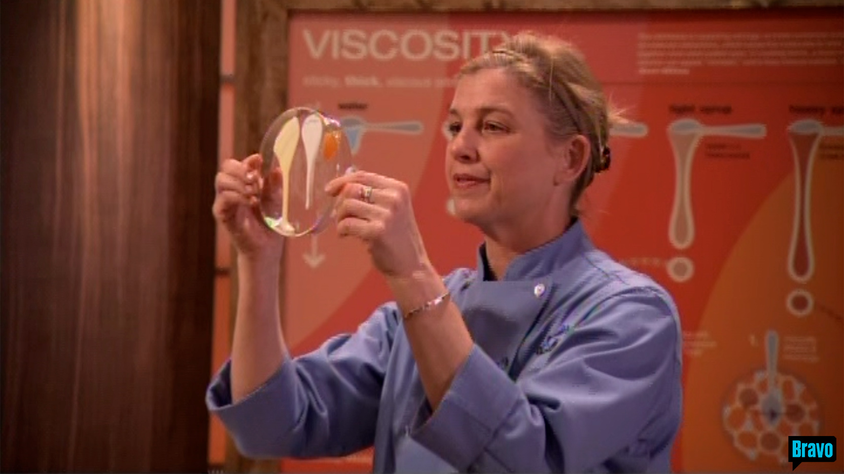 Zoom in on Chef Mary Sue Milliken&rsquo;s upper body, dressed in a blue chef top, 
                                                                                holding up a glass plate on which different thick liquids pour slowly down, 
                                                                                while standing in front of the large VISCOSITY illustration framed in stained pine, 
                                                                                on the set of Bravo TV&rsquo;s Top Chef Masters &ldquo;Blinded Me with Science&rdquo; episode.