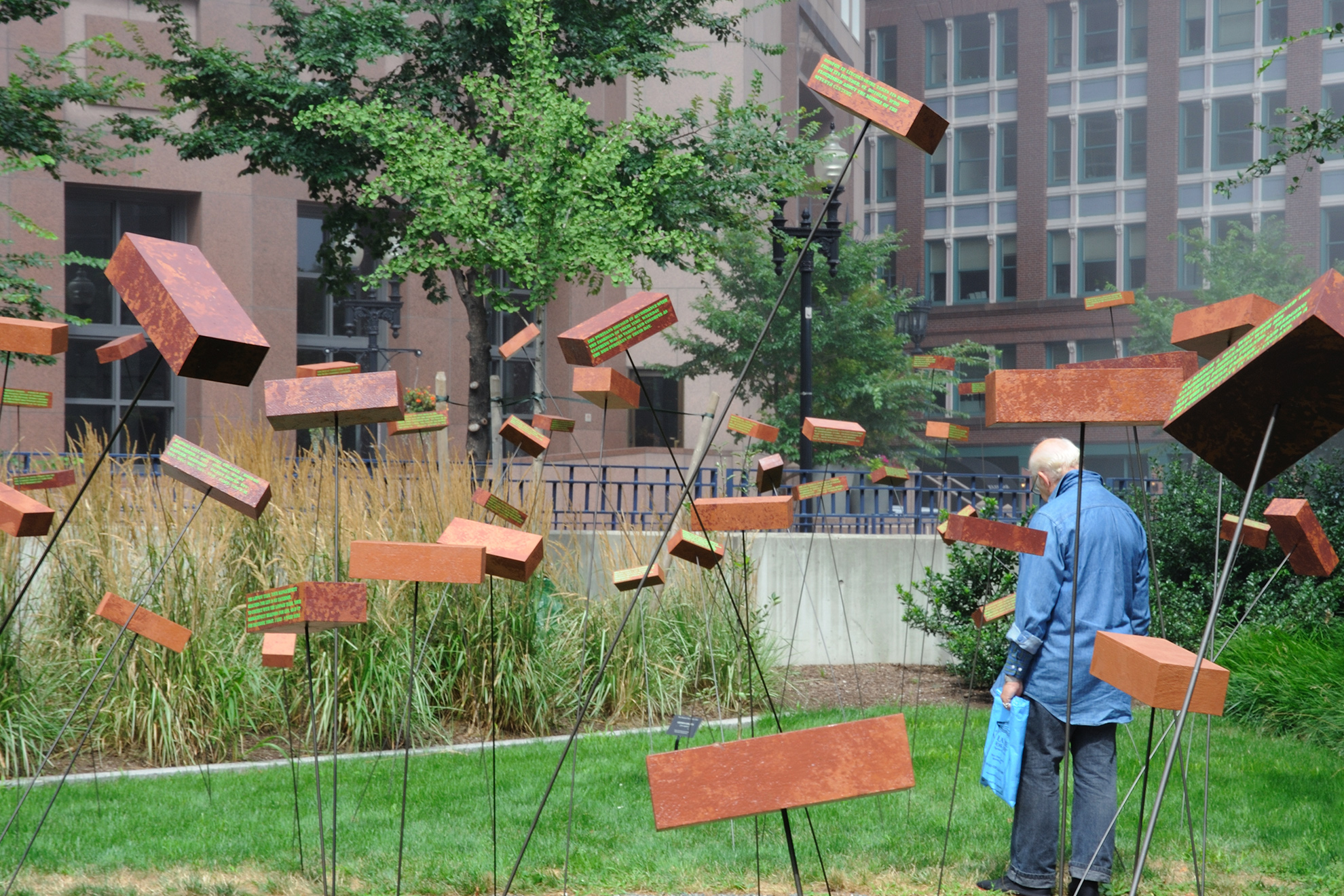 In a lush green park with bricks seemingly floating in the air at various heights, an old man holding a plastic bag reads the text painted on one of the bricks, while standing facing away from the camera.