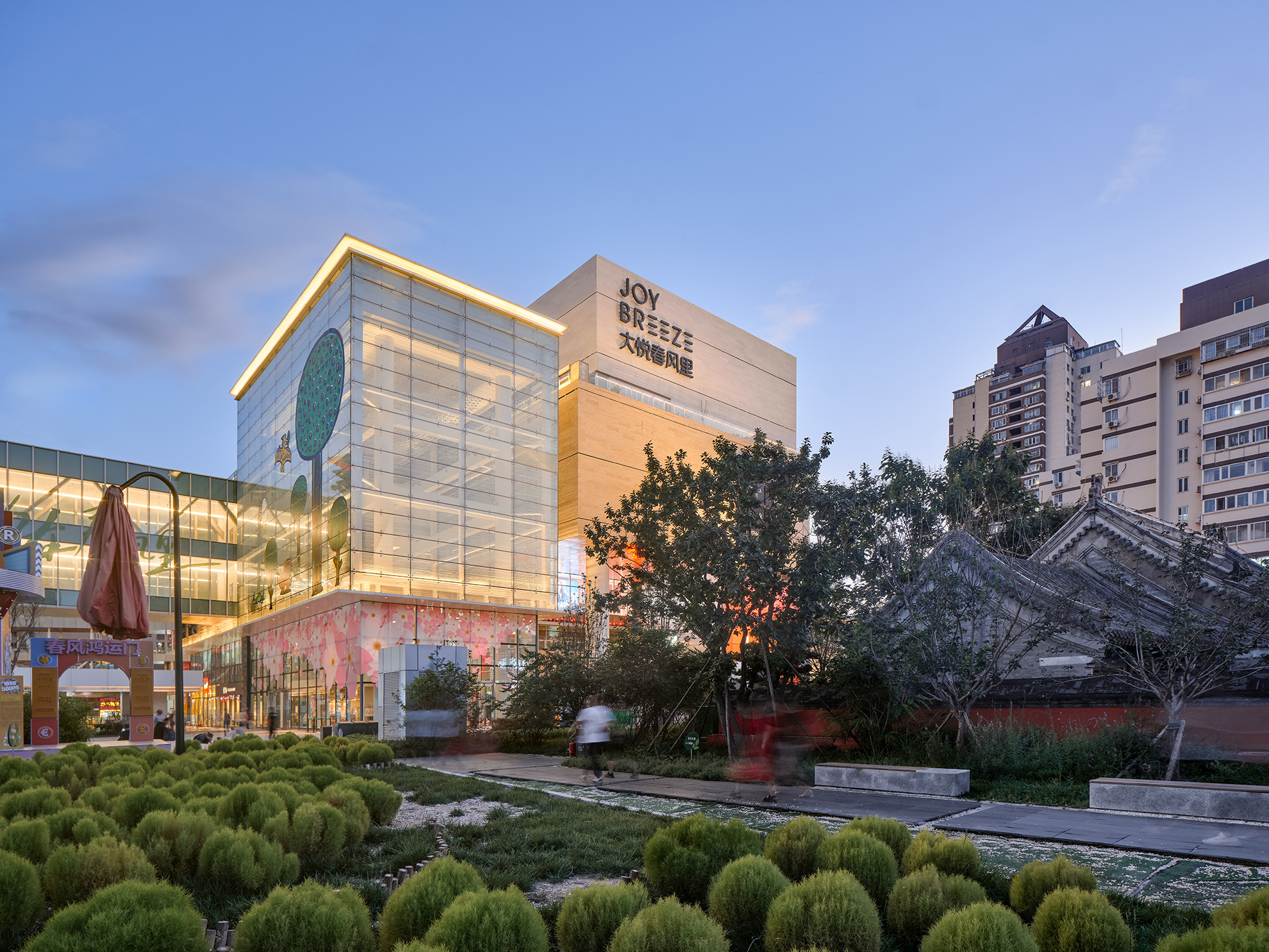 Tall glowing glass volume with glowing glass bridge on its left and stone volumes 
                                        on its right seen in the distance beyond round plant forms in the foreground and a historic temple on the right.