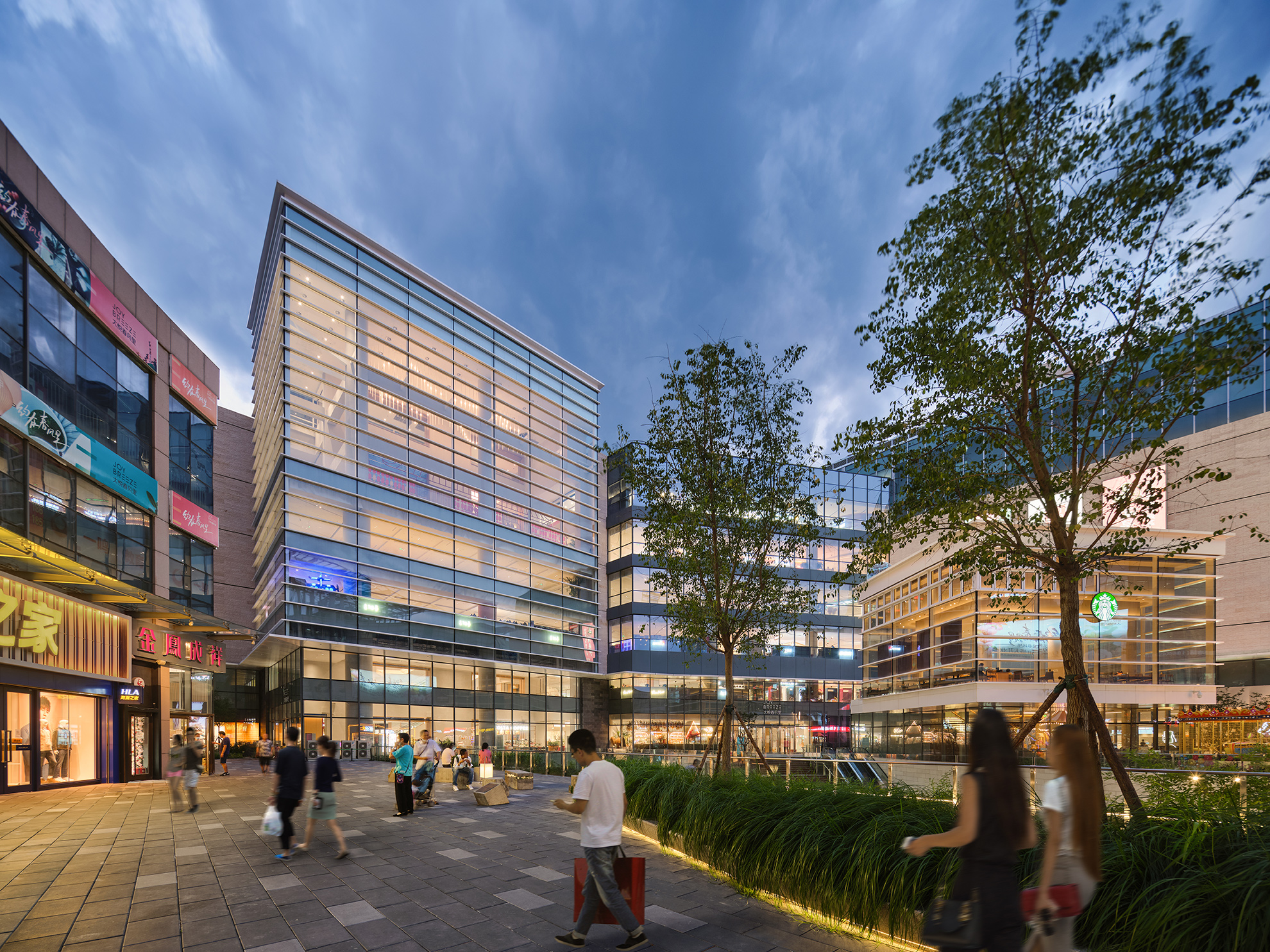 Interior courtyard of mixed-use center framed by 3-7 story &ldquo;boxes&rdquo; of glass and light beige stone, filled with people and some landscaped elements like trees and grasses, glowing-renov in the twilight hours.
