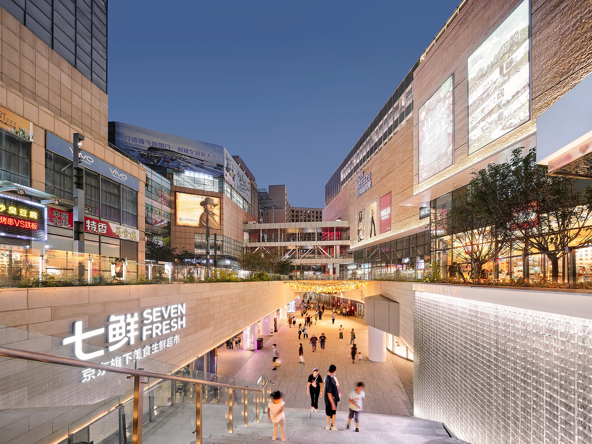 Long view, centered at the ground level, into the slow descent into the basement level retail area which is open to the sky. 
                                                                    The backlight glass wall along the grand staircase down glows bright white, suggestive of a waterfall. 
                                                                    The center is clad in a light beige stone which is lit to glow at twilight.