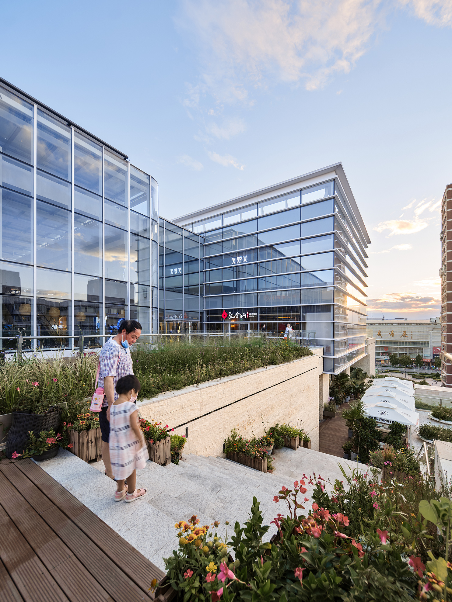 A man holds a toddler girl&rsquo;s hand, standing at the top of a stone staircase, with green planters and trees all around, and against a backdrop of glass volumes that rise up several stories.