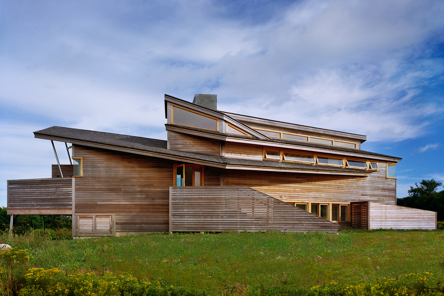 A small to medium sized house with layered angled walls and angled roofs, 
                                                                                              clad in yet unweathered horizontal cedar siding, sits in a grassy field against a big blue sky with clouds.