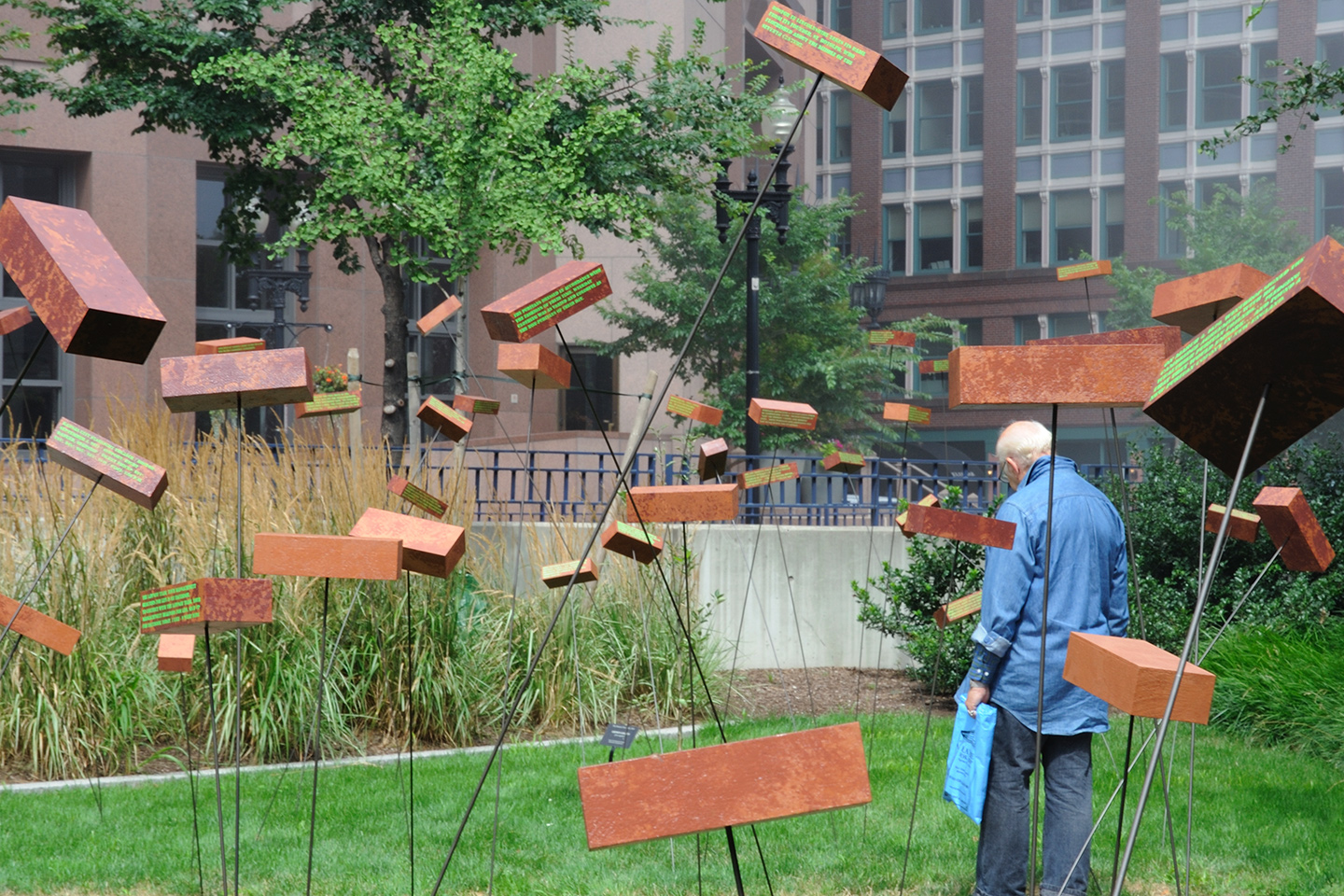 In a lush green park with bricks seemingly floating in the air at various heights, an old man holding a plastic bag reads the text painted on one of the bricks, while standing facing away from the camera.