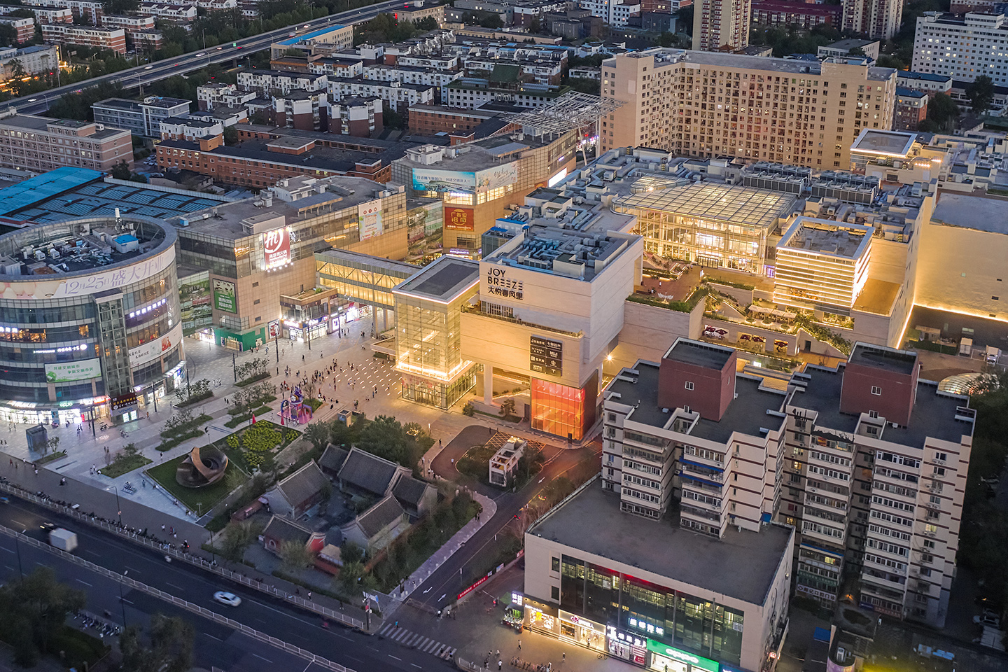 Aerial view of the large conglomerate mixed-use center, with terraces at many levels and large glass “lanterns” at many levels throughout 
                                                                            – all glass spaces and/or skylights – as well as several glass bridges, which glow to brighten and modernize the old heavy shopping center.
                                                                            In the lower right corner is overlapped a photo showing the heavy pink concrete shopping center before renovation.