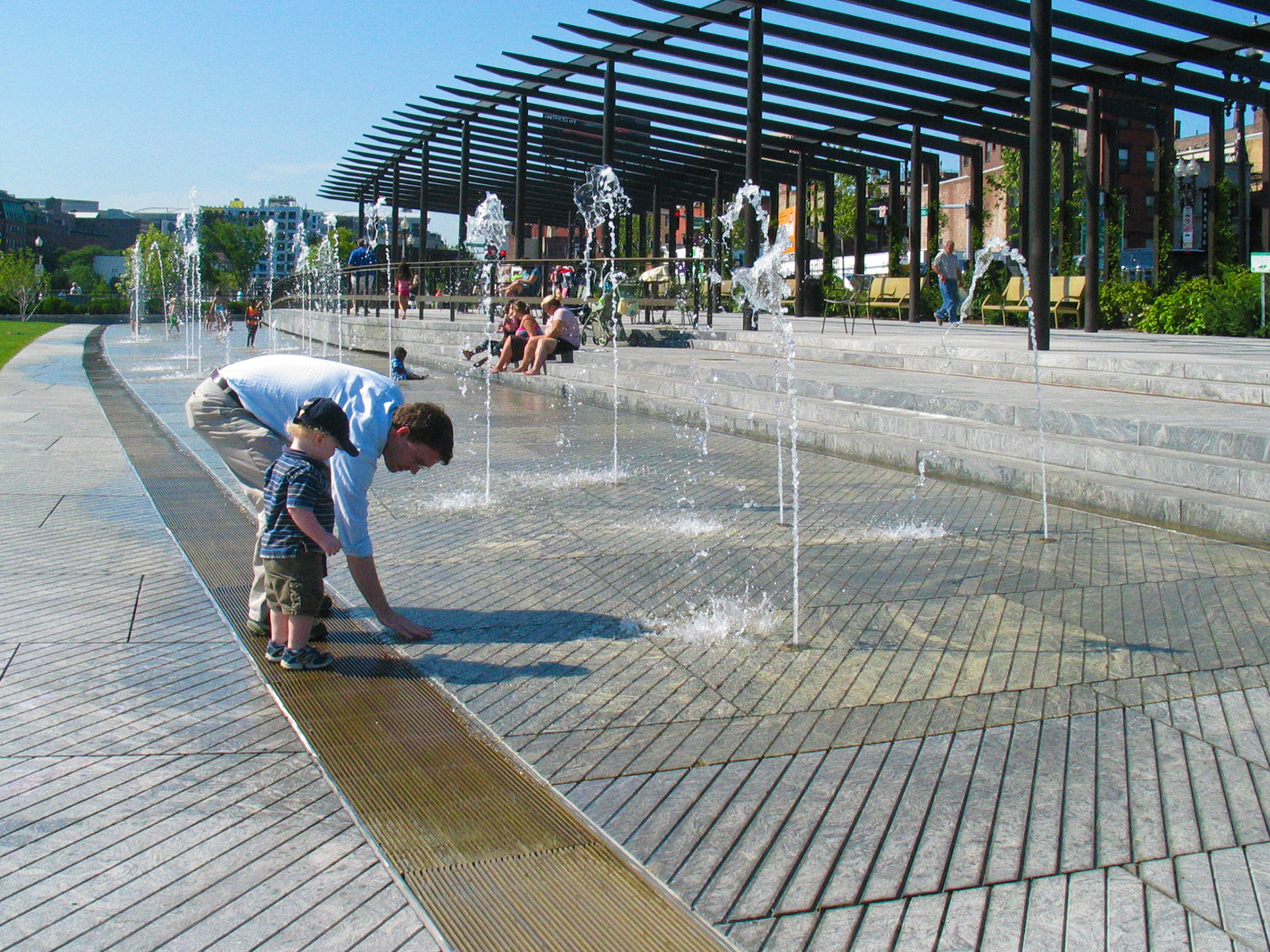 Father next to toddler standing on golden trench drain grate in the long, walk-in fountain parallel to and below the pergola zone, bends down to touch water. 
                                                        The large paver slabs in the fountain have long diagonal groved lines on them inspired by the historic non-slip pavers in the North End.