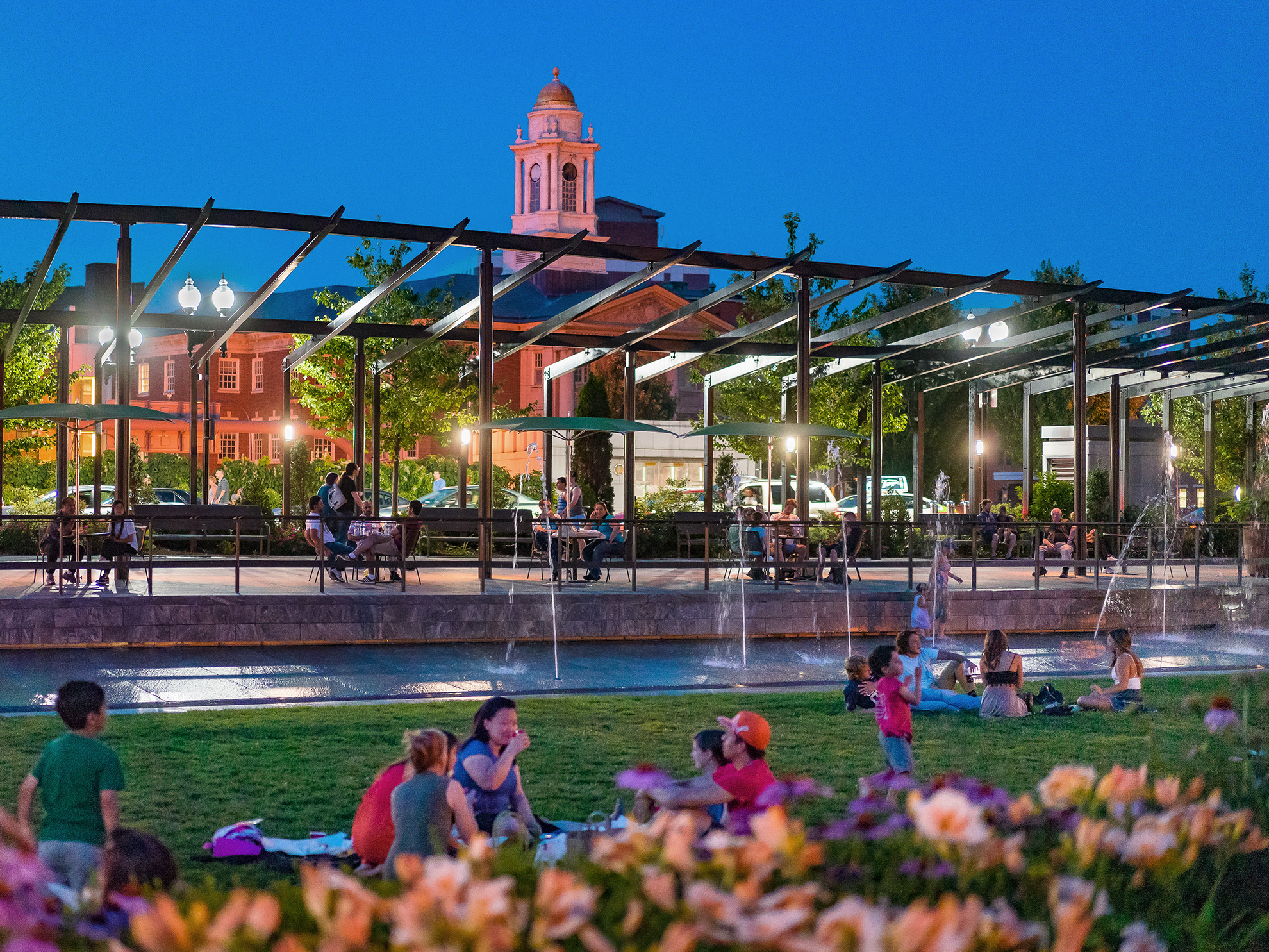 People hanging out at twilight in the parks: on the grass, in the walk-in fountain beyond, and in the space framed by the large brown painted thin steel pergolas further back. 
                                                            The buildings of the North End can be seen through the pergola.
