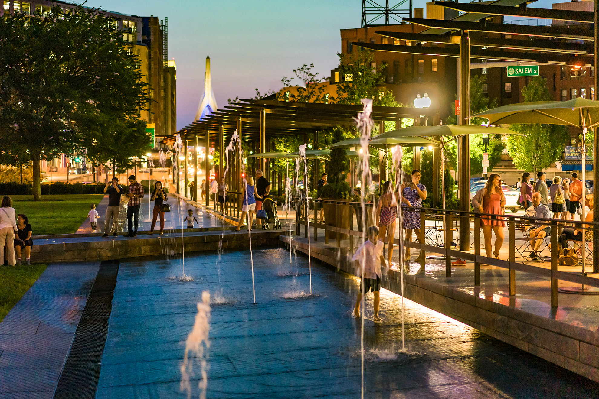 Colorful summer twilight in the park, with people walking and hanging out in social groups everywhere: 
                                                    on the grass, on the slightly elevated walk above the fountain zone which leads pedestrians into the space under the pergolas, 
                                                    and in the podium area under the pergolas. A few kids play in the walk in fountain as jets of water shoot up.