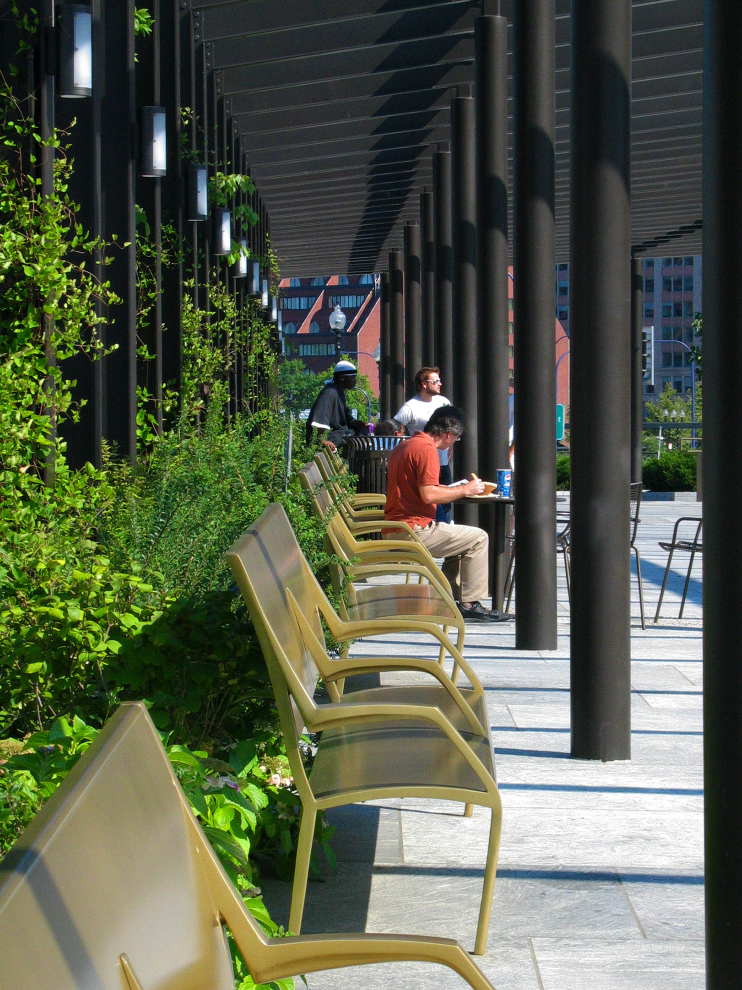 Man in a red shirt sits in a row of elegantly angled golden benches, writing at a small round table he pulled close. 
                                                            The row of benches sits between a row of dark brown steel columns
                                                            and a row of dark brown steel purlins, which are projecting out of the green bushes behind.