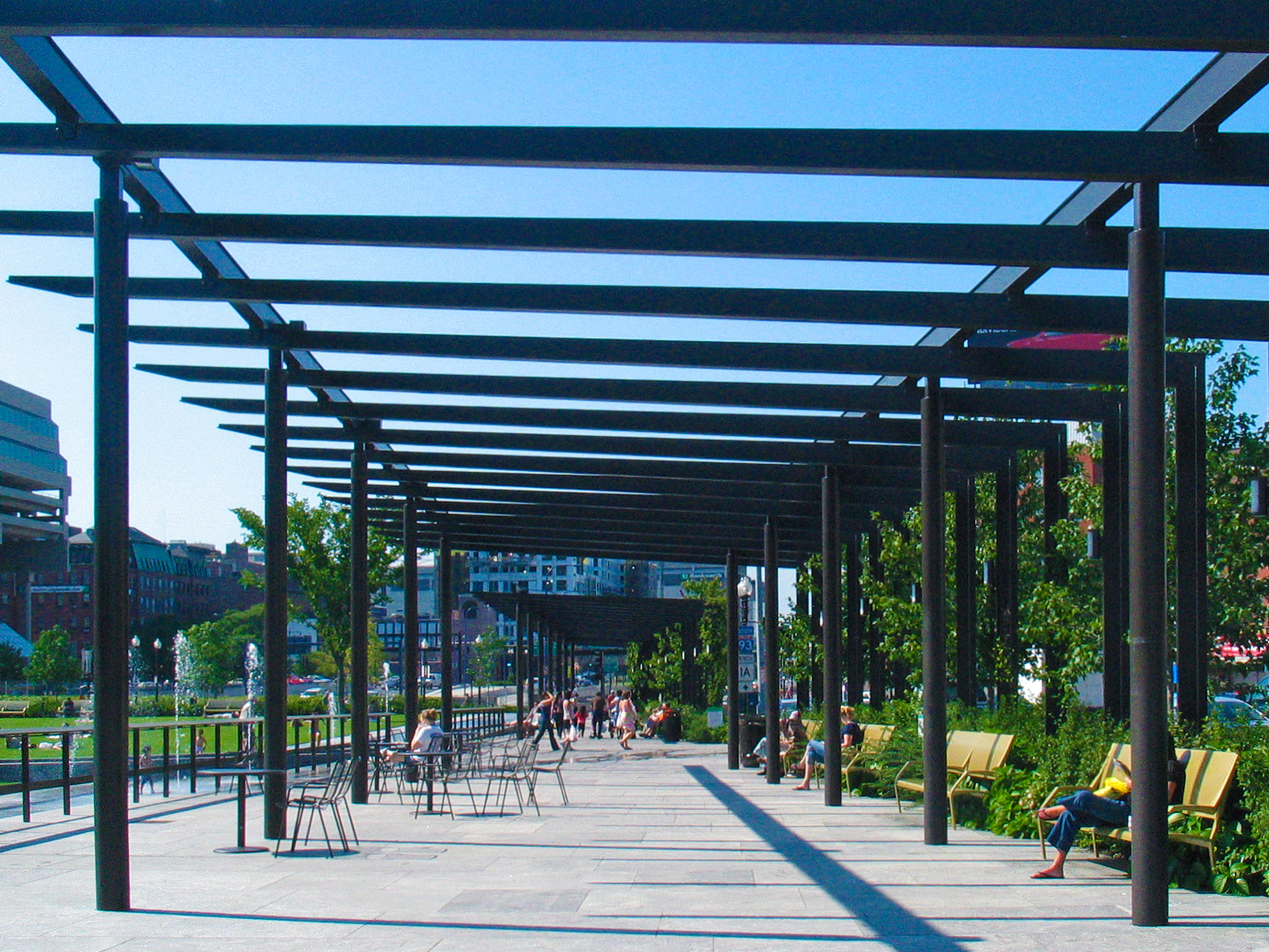 Tunnel view through all three steel pergola painted dark brown, highlighting continuity and rhythm of thin structural members.
                                                        Some people are walking within the wide, semi-covered space paved with light grey stone, while others are sitting on metal benches and at metal tables.