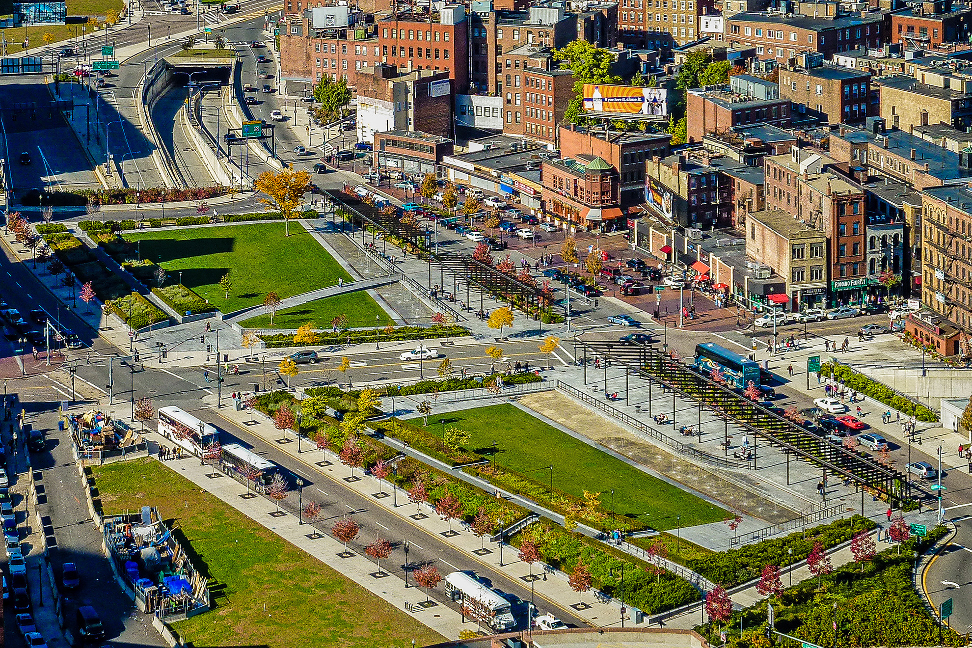 Aerial view of North End Parks, set between two large roads and Boston&rsquo;s red-brick clad North End, with two highways coming out from under them and headed underground again.
                                                        From south-west to north east, the parks geometry shows bands/zones of: trees, bushes and plants, green lawns, walk-in fountains, dark brown steel pergolas covering a very wide walkway, 
                                                        and plants again.