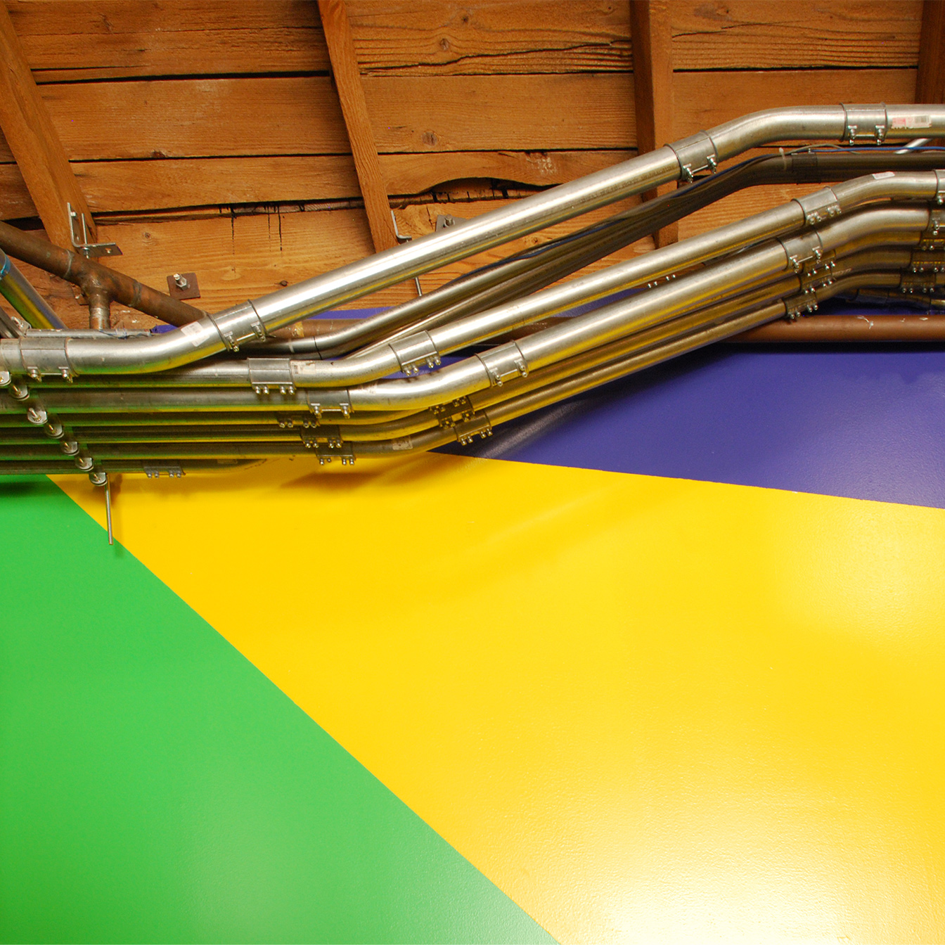 Zoom in on many electrical conduits in metal pipes running between the slanted wood roof-ceiling and the painted wall below, triangulated into areas of green, yellow, and blue.