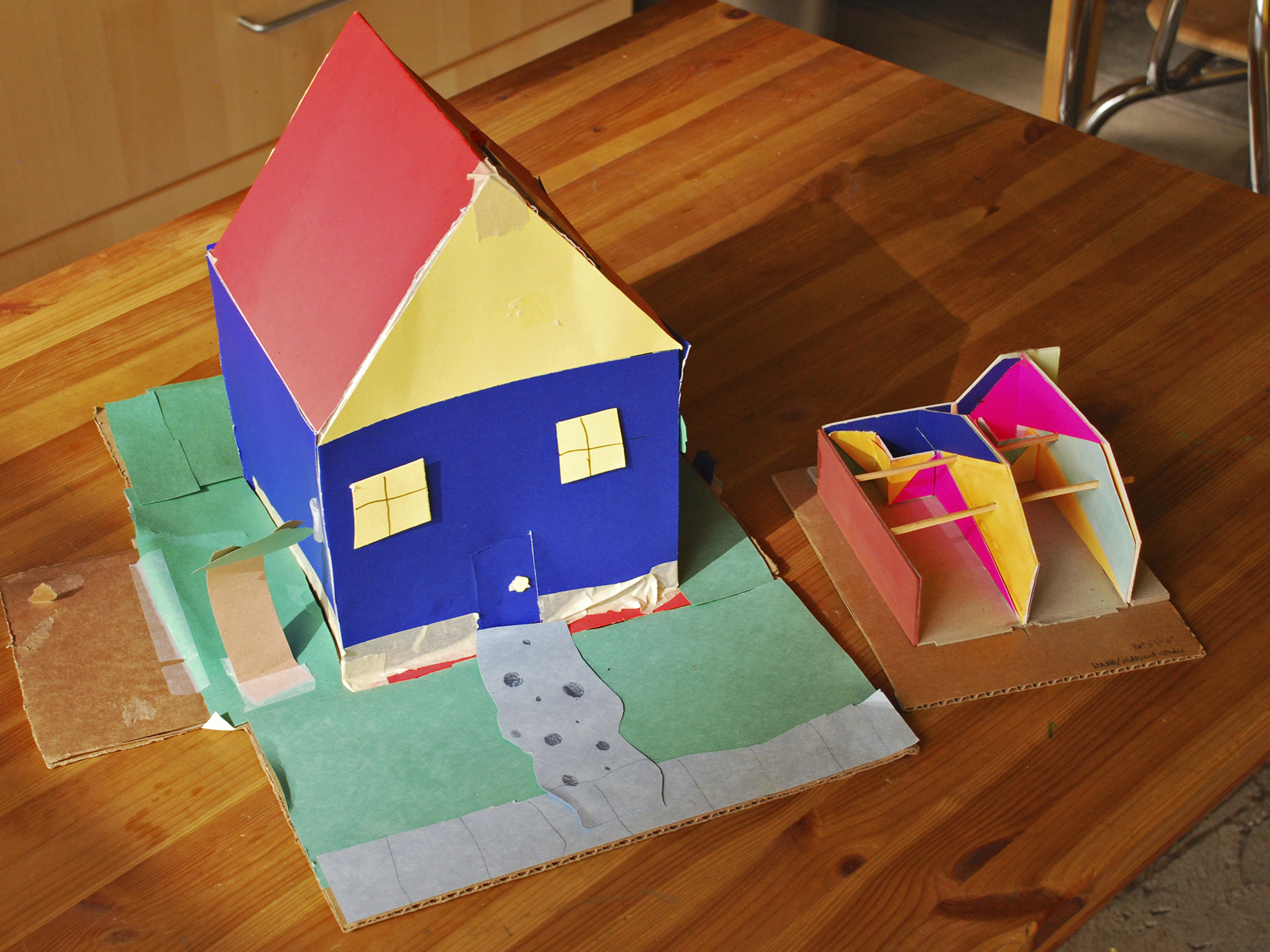 A large child&rsquo;s colorful paper model of a house, in red, yellow, and dark blue, next to the small model of the studio paint design.