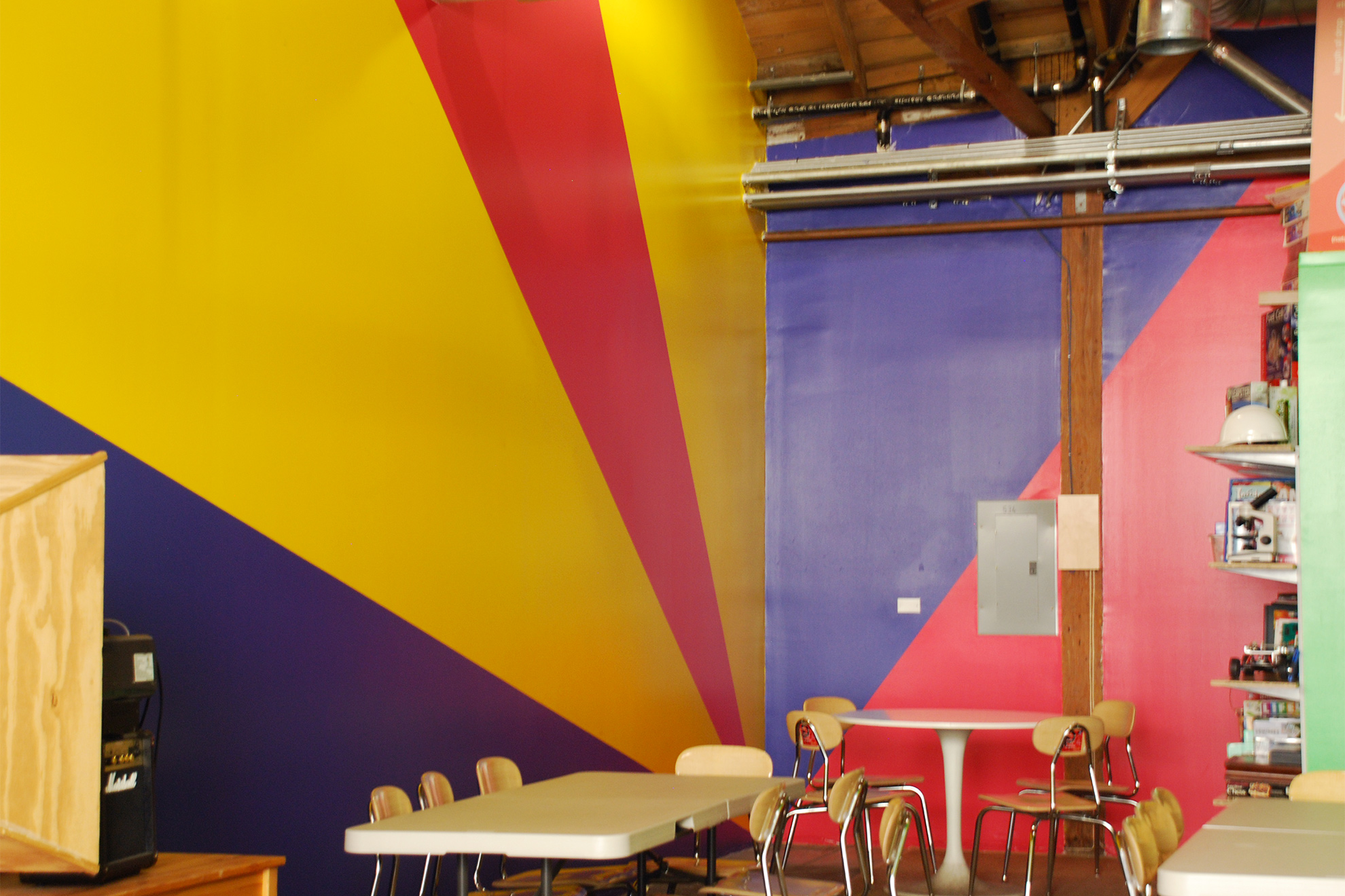 Left back corner of one of the workshop spaces where triangular painted areas meet in the bottom corner.
                                                            On the left wall, from bottom of the wall up are triangular painted areas in blue, yellow, red, yellow.
                                                            On the back wall, intersected by a wood pilaster, is a red painted area on the bottom and right, and a blue painted area on the left and top.