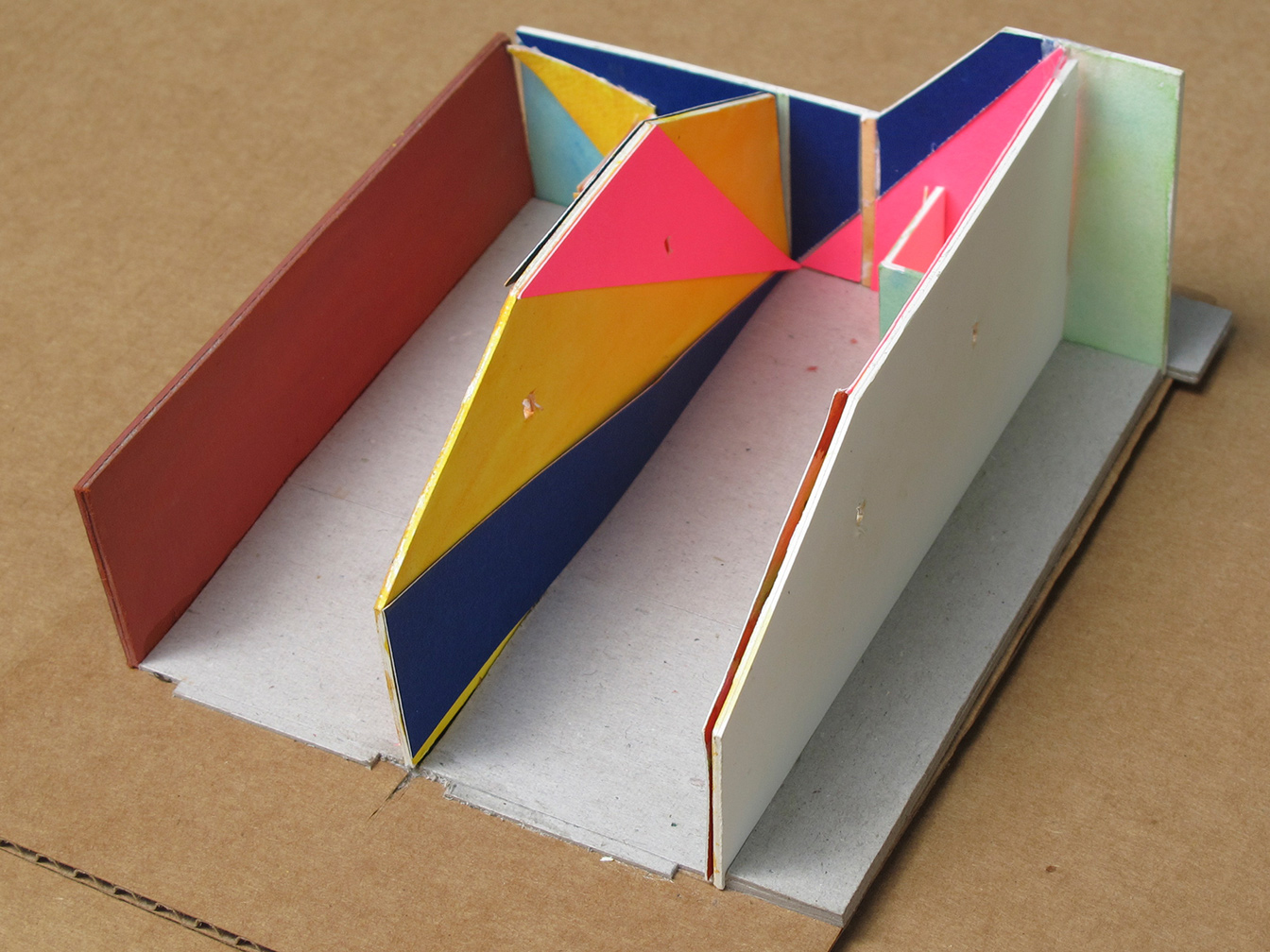 Side view into cardboard model shows brick wall in one space, and painted wall in the other, with triangular areas in blue, yellow, and red expanding toward the front.