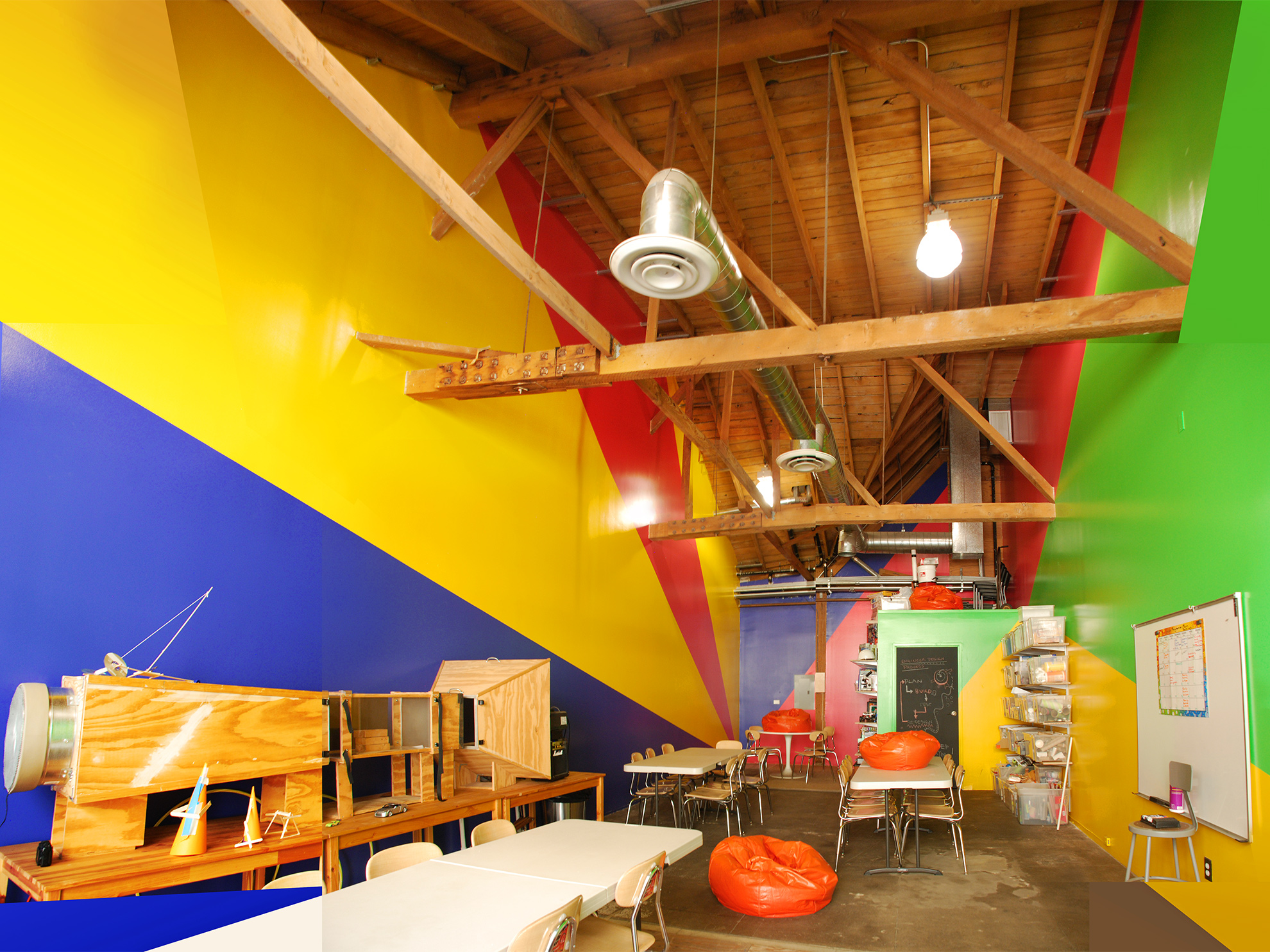 20 ft tall workshop interior with walls colored in angular shapes, mimicking a perspective emanating from the back wall out. 
                                                            Paint colors are red and blue on the back wall; reddish-magenta, blue, and yellow on the left wall; 
                                                            and yellow and green on the right wall and on the short bathroom wall which pops out into the space.
                                                            The ceiling, which angles up and down in complex directions, is of exposed wood plank and framing,
                                                            as are the large beams which cross the space above.