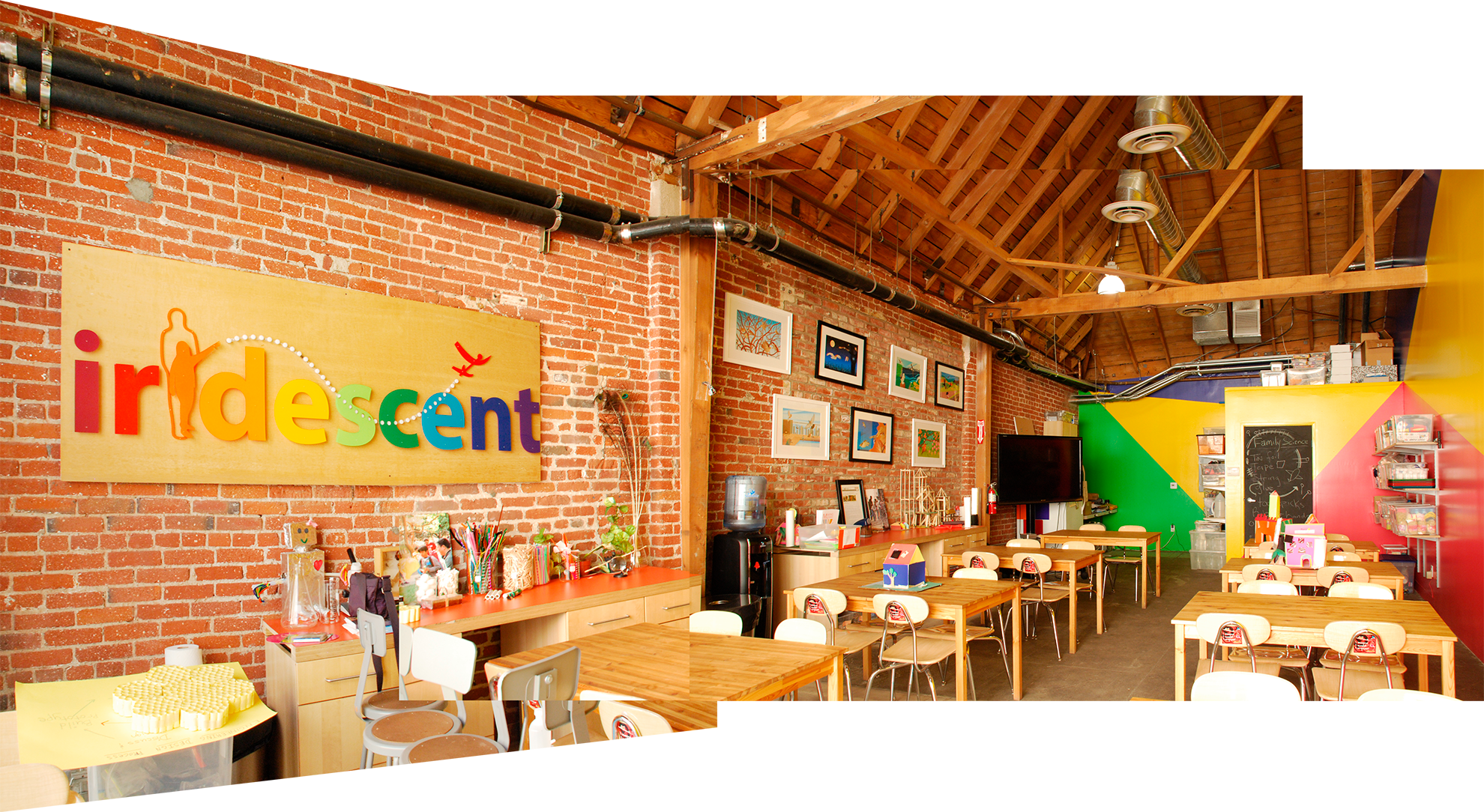 Interior of children's classroom with red brick walls on left, warm wood angled ceiling-roof, and colorful triangulated areas of color on back and side walls in green, blue, yellow, and red. 
                A big rainbow colored Iridescent logo hangs on the brick wall, together with some framed art. The tables and chairs are in a light colored wood.