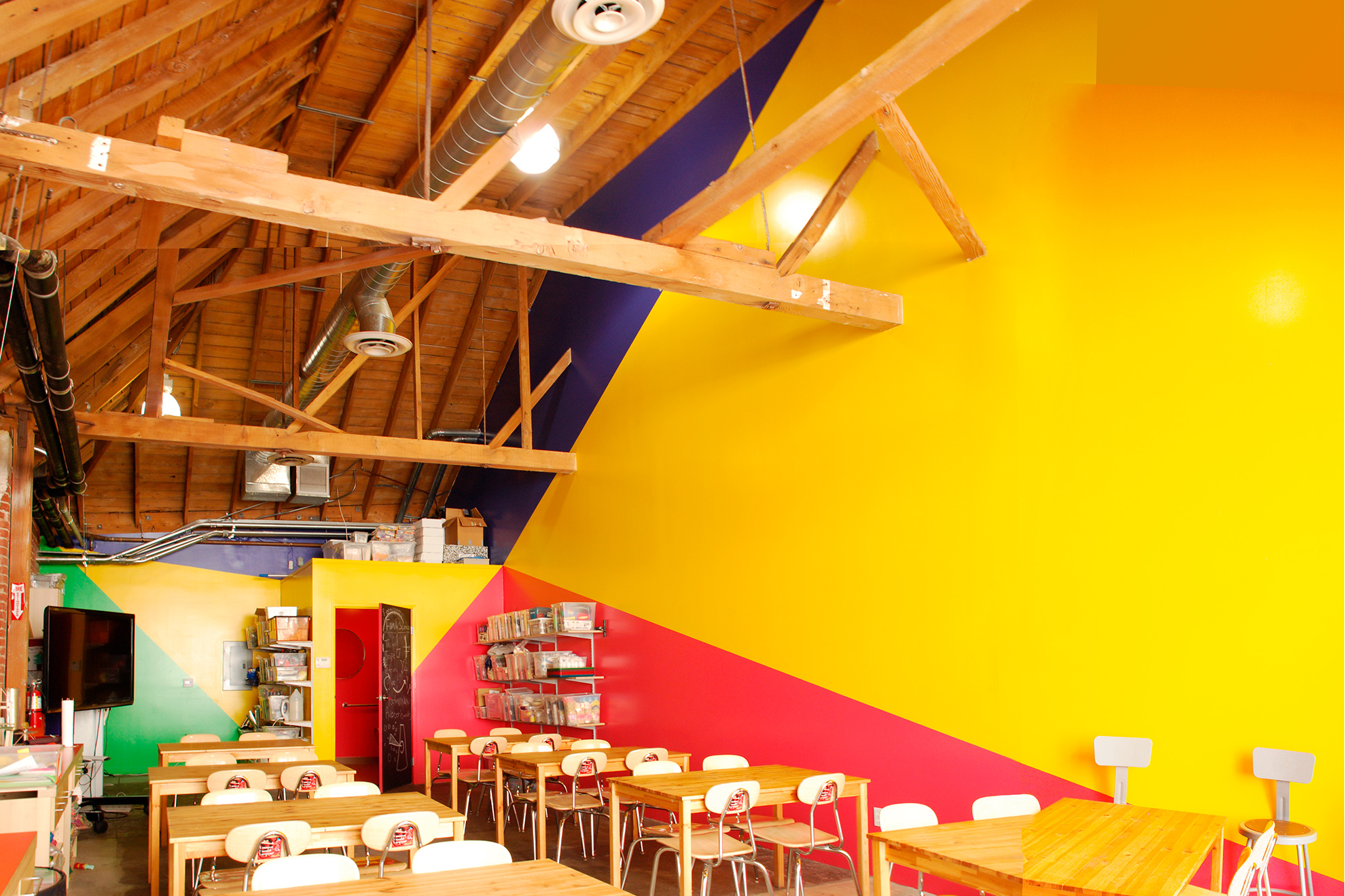 Interior of classroom focusing on triangulated painted back and right side walls, in reddish-magenta yellow (the most prominent), green and blue.