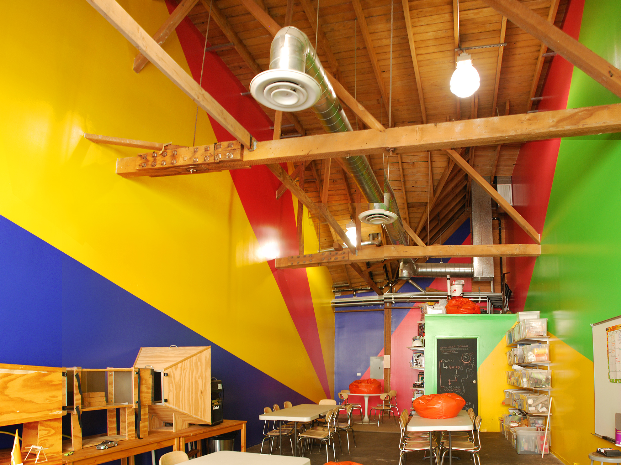 20 ft tall workshop interior with walls colored in angular shapes, mimicking a perspective emanating from the back wall out. 
                                                                Paint colors are red and blue on the back wall; red, blue, and yellow on the left wall; 
                                                                and yellow and green on the right wall and on the short bathroom wall which pops out into the space.
                                                                The ceiling, which angles up and down in complex directions, is of exposed wood plank and framing,
                                                                as are the large beams which cross the space above.